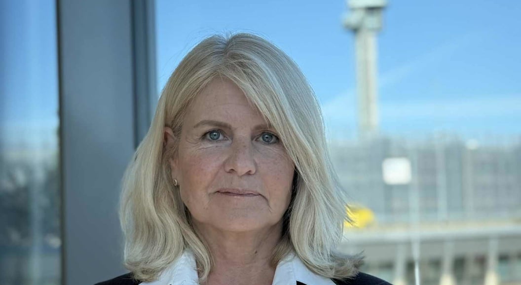 Woman in a blazer with a white shirt stands in front of a glass window overlooking an airport terminal and control tower in the background.