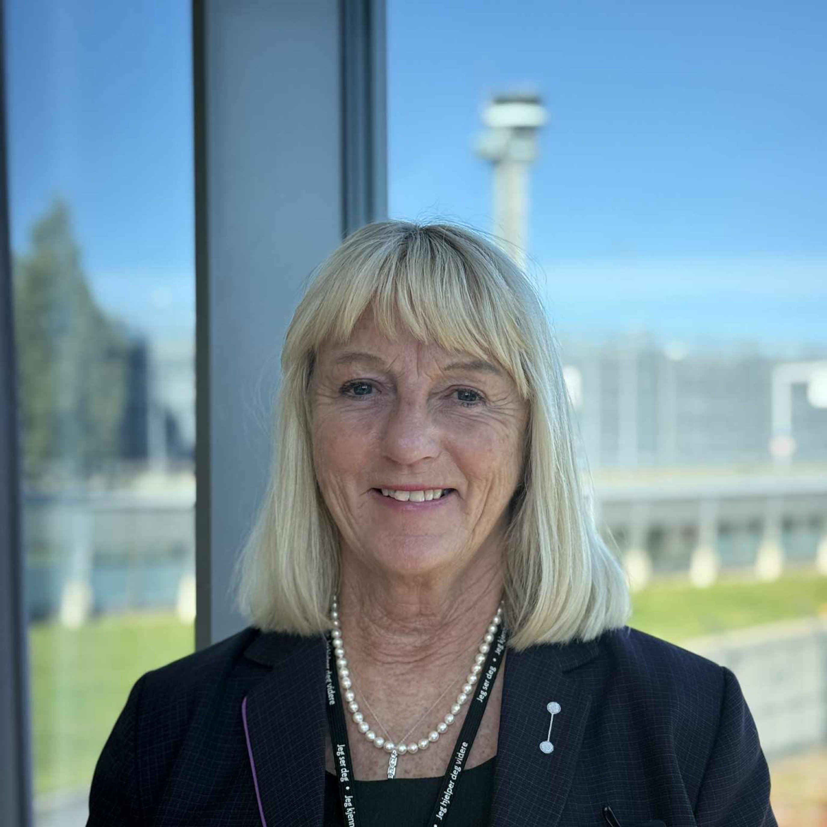 Woman with blonde hair in a dark blazer smiling in front of a window overlooking an open landscape and a blurred structure in the background.