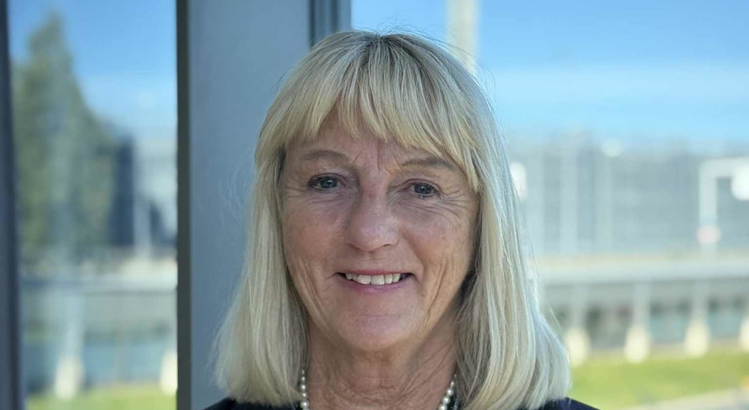 Woman with blonde hair in a dark blazer smiling in front of a window overlooking an open landscape and a blurred structure in the background.