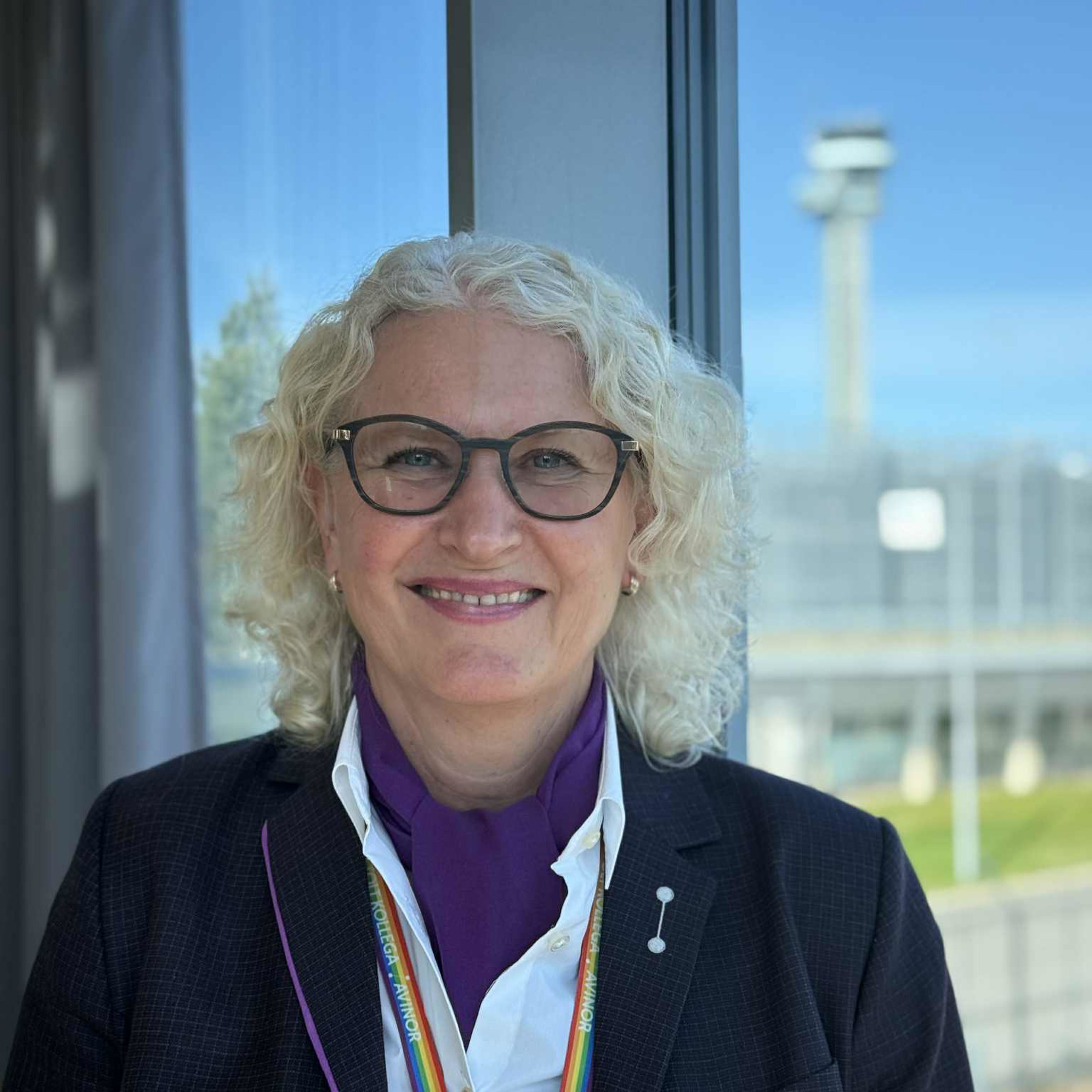 Woman with light curly hair and glasses stands by a window wearing a dark jacket and shirt, in front of an airport background with a clear blue sky.