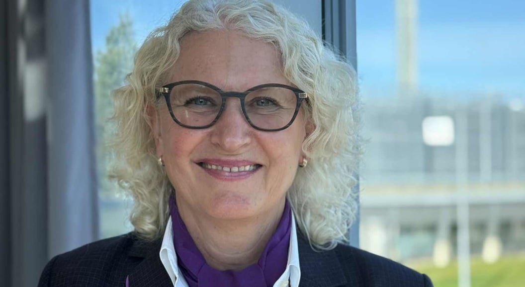 Woman with light curly hair and glasses stands by a window wearing a dark jacket and shirt, in front of an airport background with a clear blue sky.