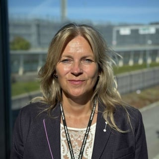 Woman in formal attire smiling outdoors with clear skies and buildings in the background.