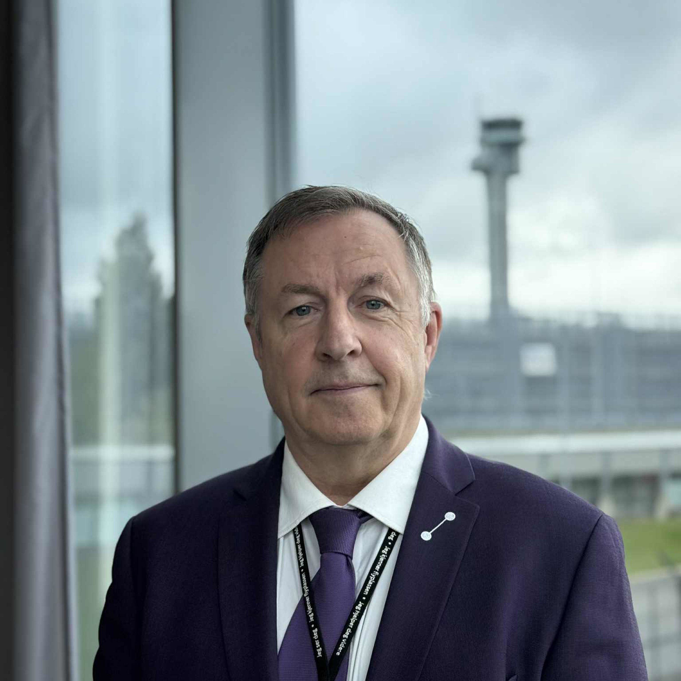 A man in a suit stands in front of a large window overlooking an airport terminal and control tower in the background.
