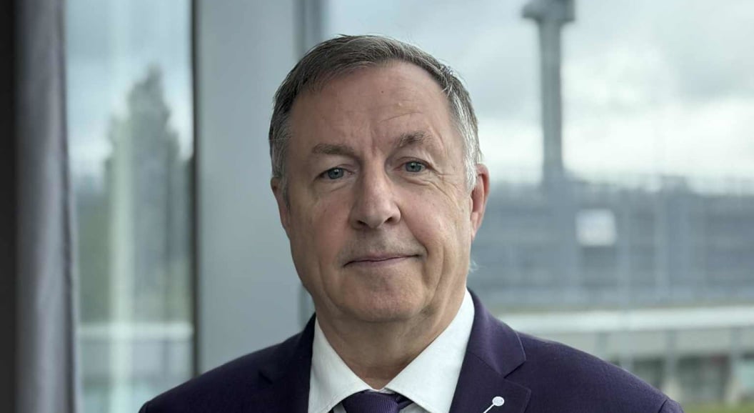 A man in a suit stands in front of a large window overlooking an airport terminal and control tower in the background.