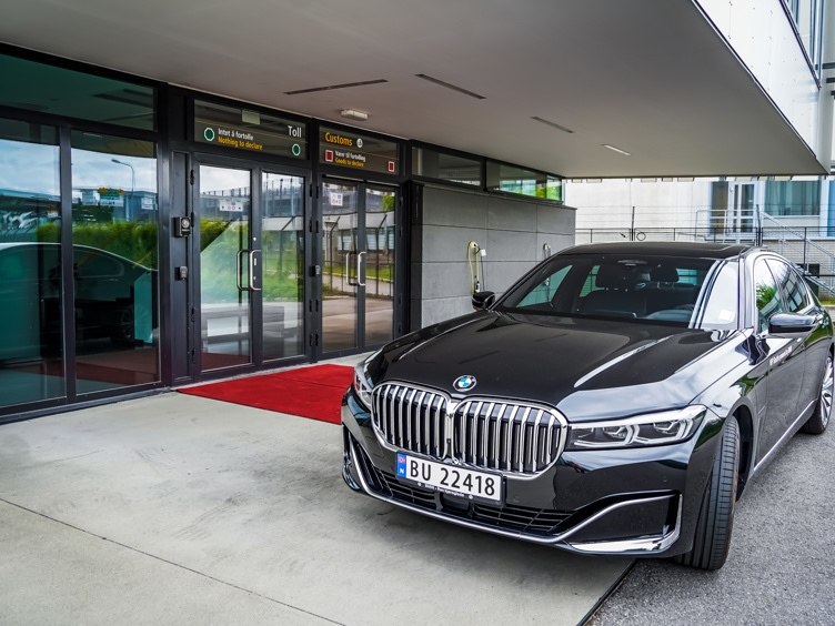 Black BMW parked in front of a customs check-in at a building with glass doors, red carpet along the entrance.