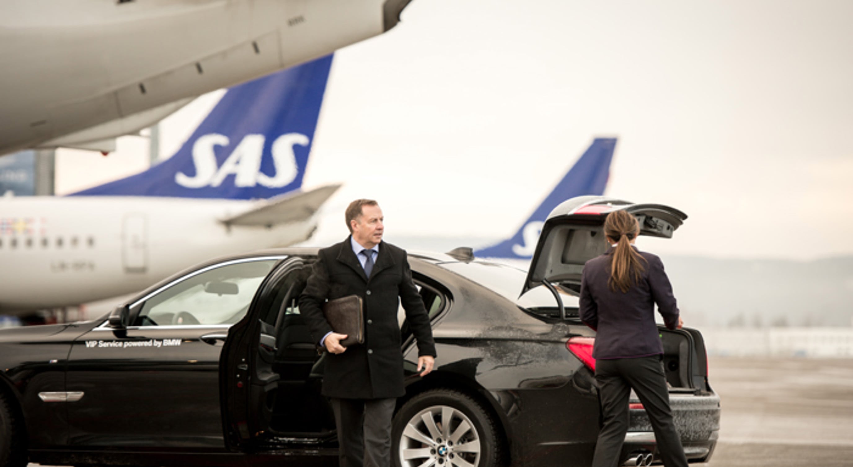 Businessman getting out of a luxury car at an airport, with SAS planes in the background. A woman is loading luggage into the car's trunk.