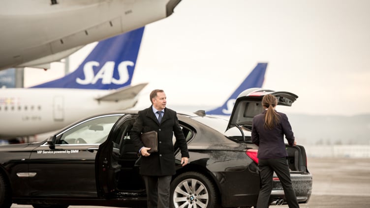 Businessman getting out of a luxury car at an airport, with SAS planes in the background. A woman is loading luggage into the car's trunk.
