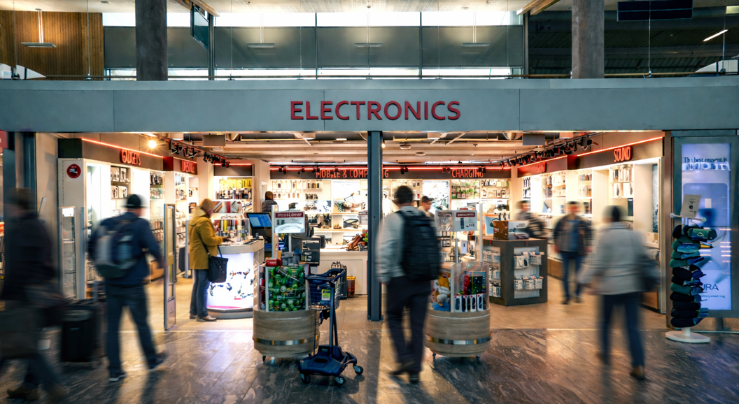 Salesperson assisting a customer at a CAPI store counter in an airport, surrounded by tech products and colorful shelves