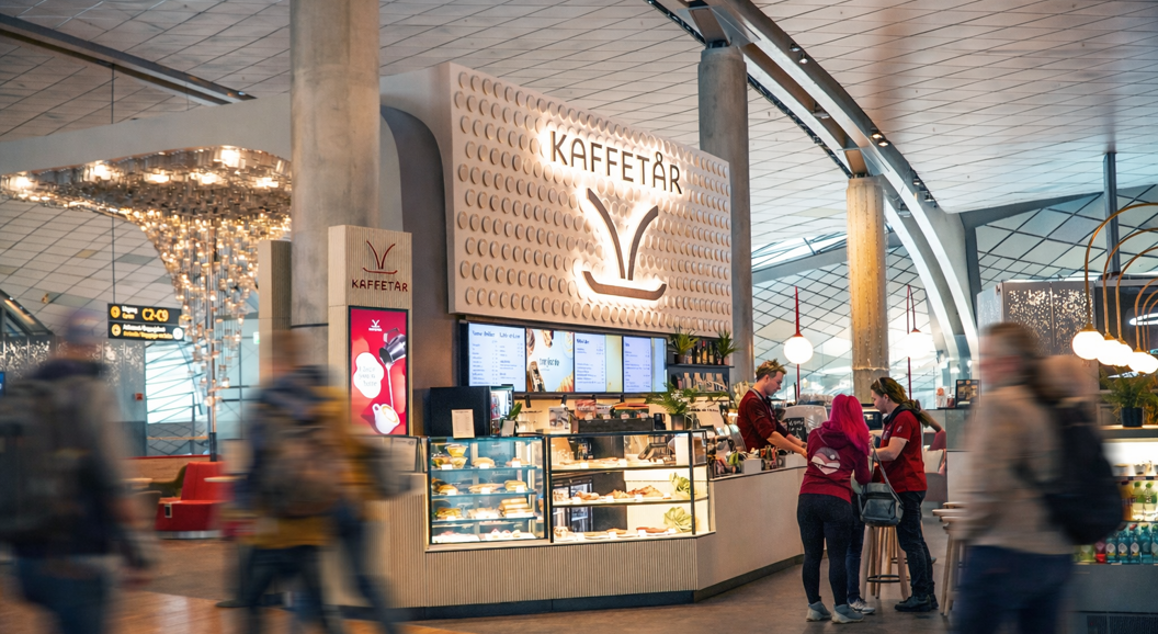 Norwegian airport lounge with seating area, decorative wall design with 'Norge', directional signs for gates and toilets, and passengers working on laptops