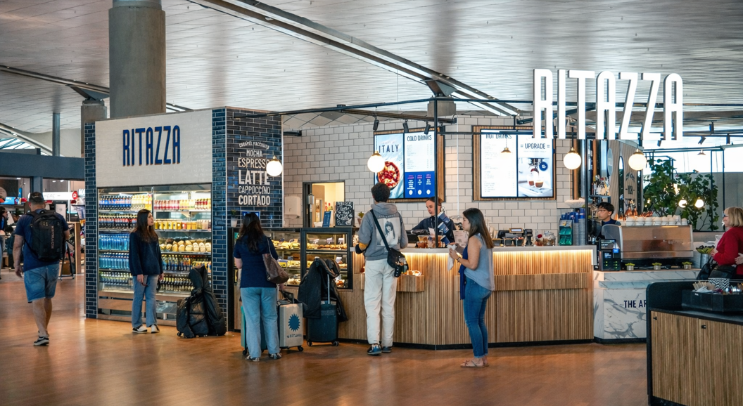 Blue Ritazza cafe sign surrounded by greenery, with a reflection of a modern interior in the glass window, suggesting a cozy and inviting atmosphere