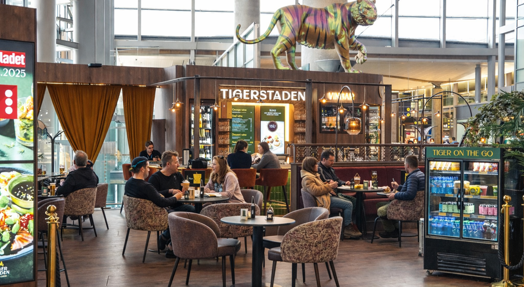 Modern airport bar and lounge area with seating, featuring a large decorative tiger sculpture above "Tigerstaden Vinbar" sign, with patrons and staff visible, and a directional sign for gates E, F, G in the background