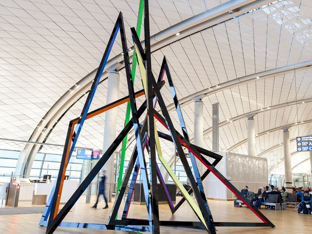 Moderne skulptur med fargerike, geometriske former i en stor, lys sal med buet tak, mennesker og informasjonstavler i bakgrunnen