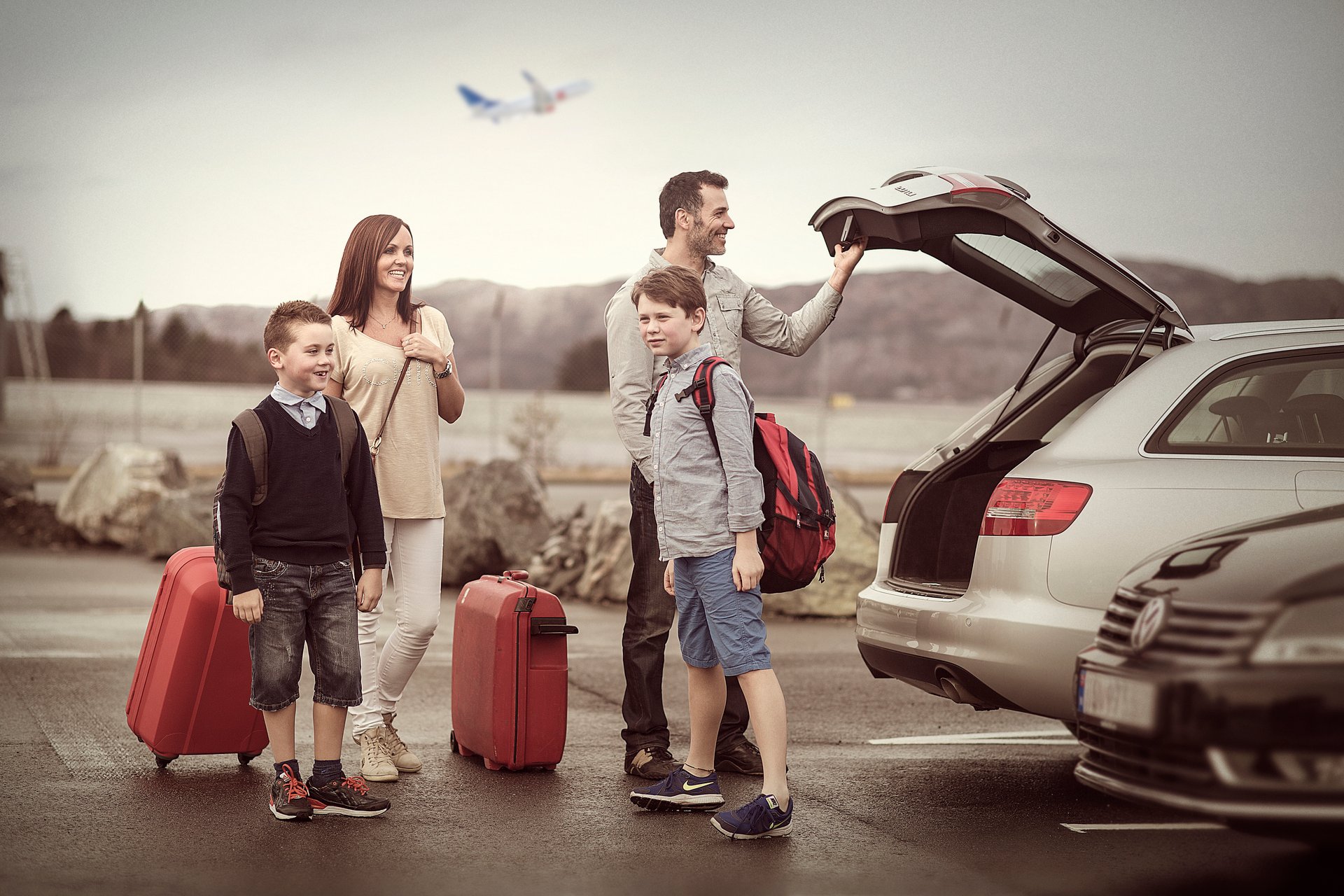 Familie ved parkeringsplass med bagasje og bil, klar for reise. Fly i bakgrunnen.