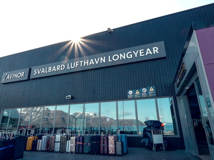 Svalbard Lufthavn Longyear airport terminal with rows of luggage carts and suitcases outside, mountains reflected in the windows, and sun flare above the building