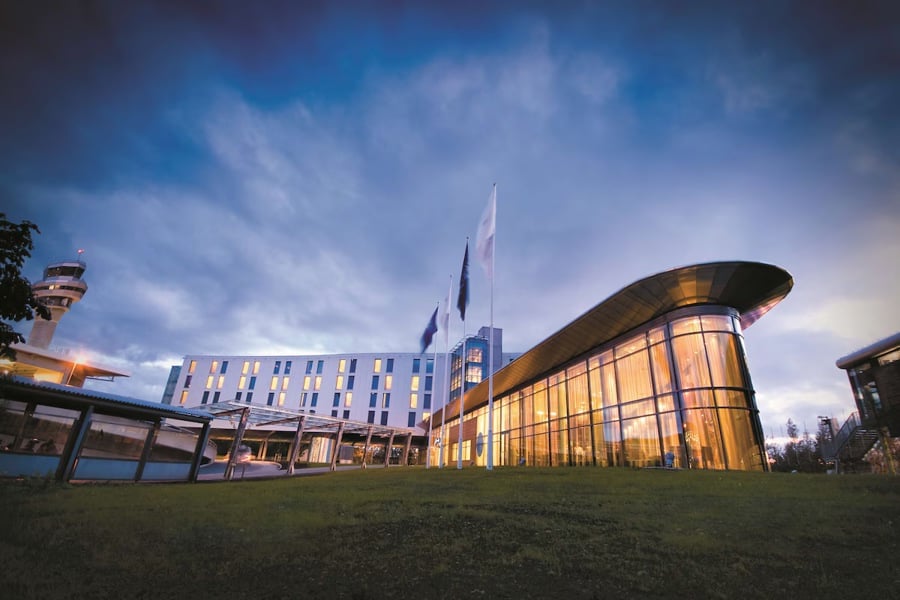 Moderne hotellbygning med glassfasade ved siden av flyplasstårn i skumringen, omgitt av grønt gress.
