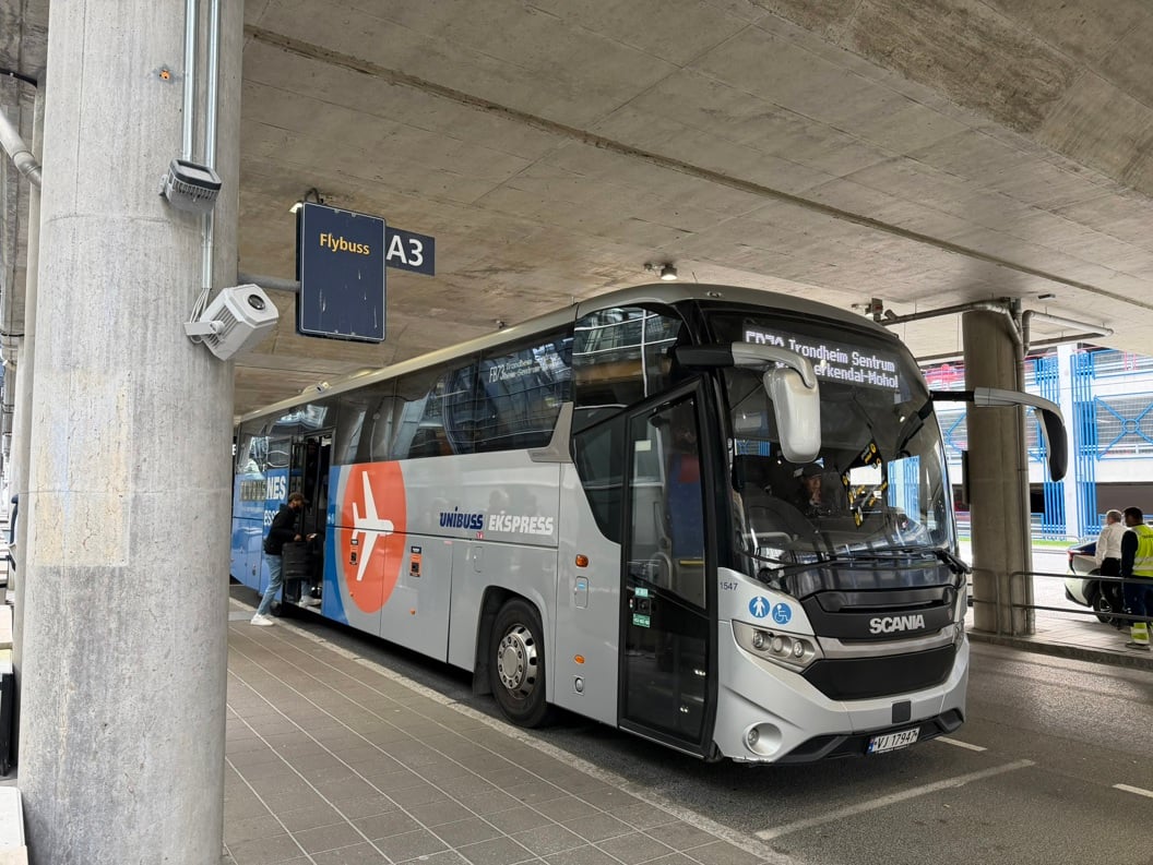 Blue bus outside an airport terminal in Norway, with passengers disembarking and some standing with luggage. Modern building and parking lot in the background. Gray sky and humid weather.