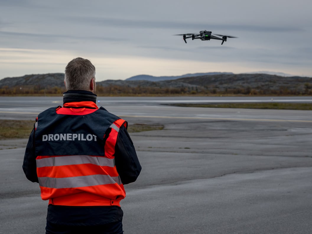 Mann i oransje jakke med dronepilot-vest som flyr drone på en flyplass