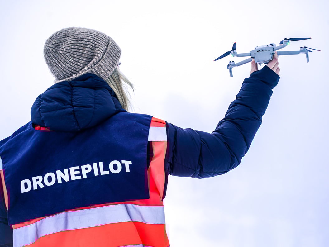 Woman in an orange jacket with a drone pilot vest holding a drone in her hand up to the sky